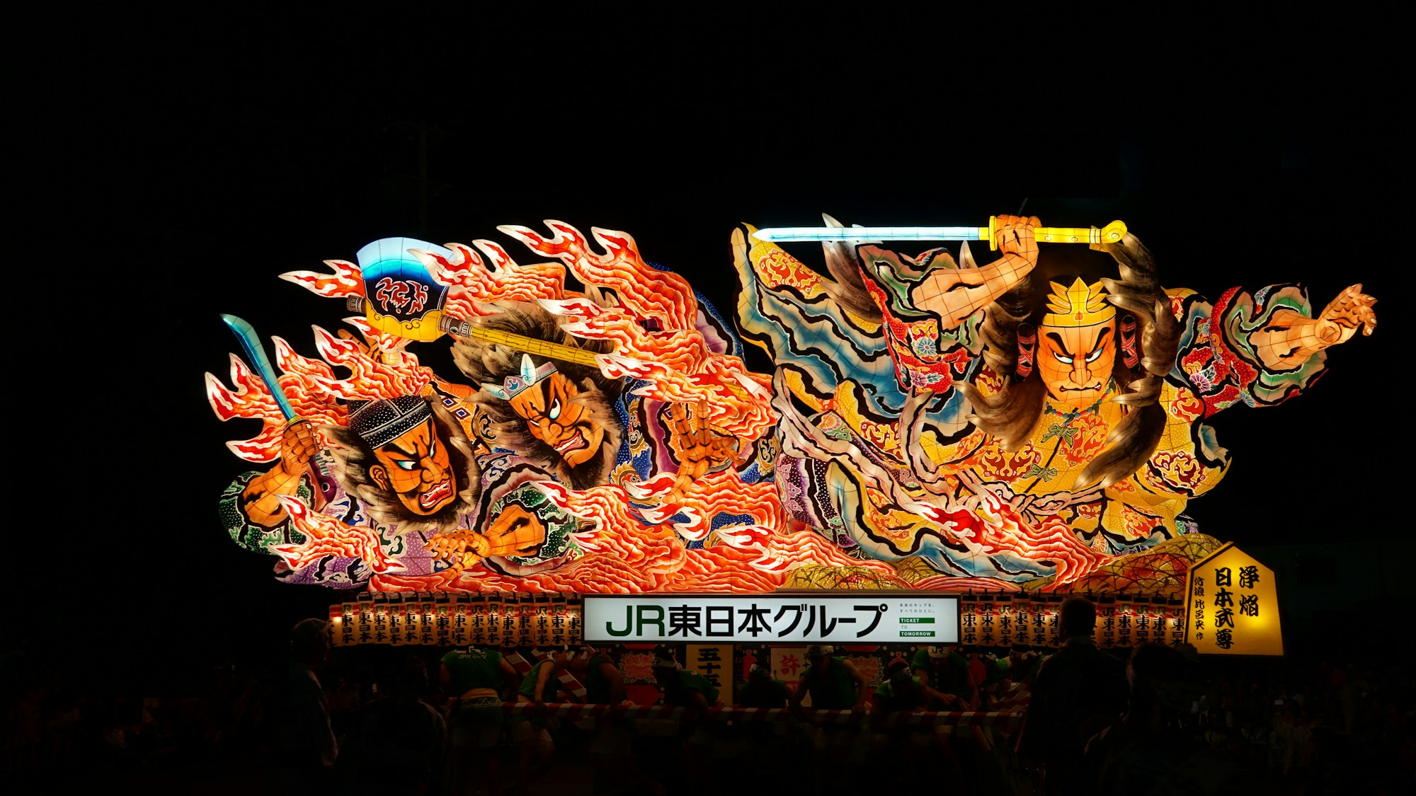 Bailarines haneto con trajes coloridos saltando frente a una carroza nebuta iluminada