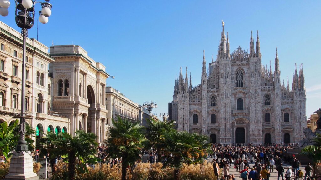 Foule de visiteurs devant le Duomo de Milan un jour d'été, sous un ciel bleu et des palmiers