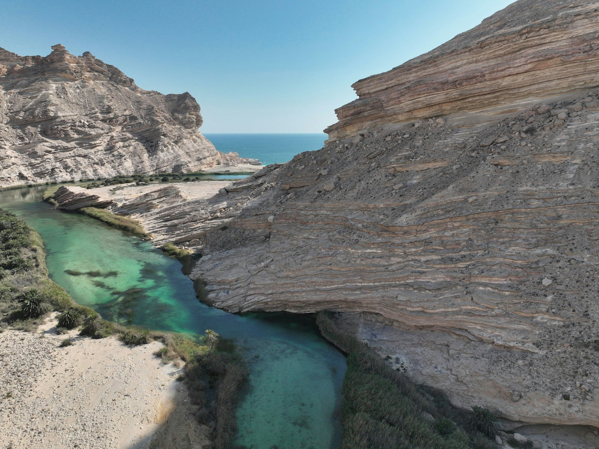 Ein türkisfarbenes Wadi, das zwischen Felsklippen ins Meer mündet, Oman