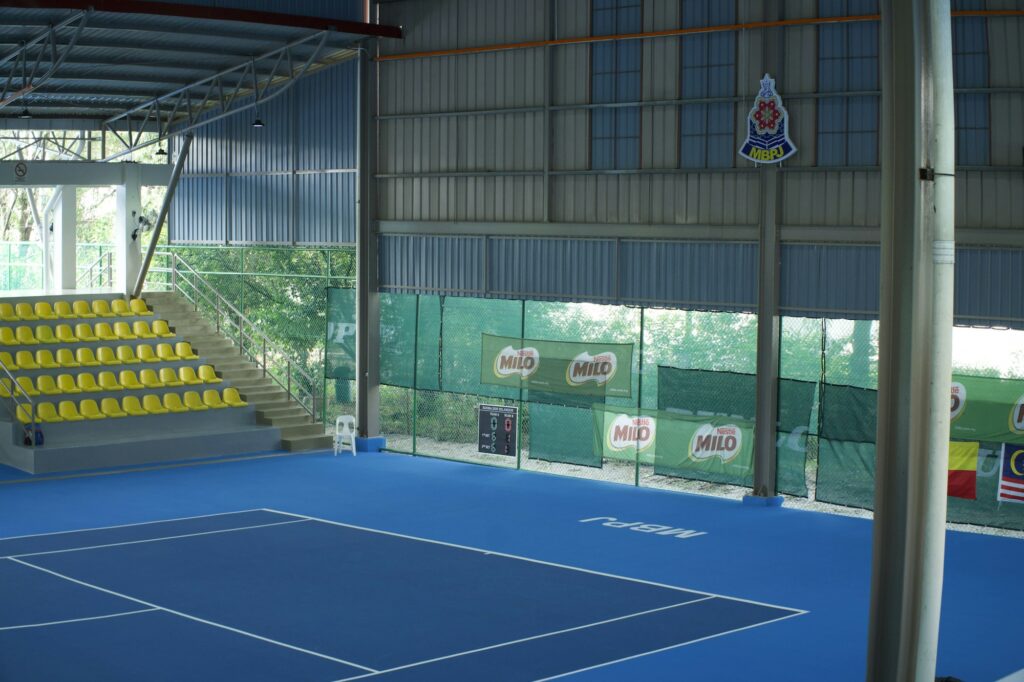 Court de tennis bleu vide et gradins jaunes sous une toiture métallique, avant un tournoi en salle