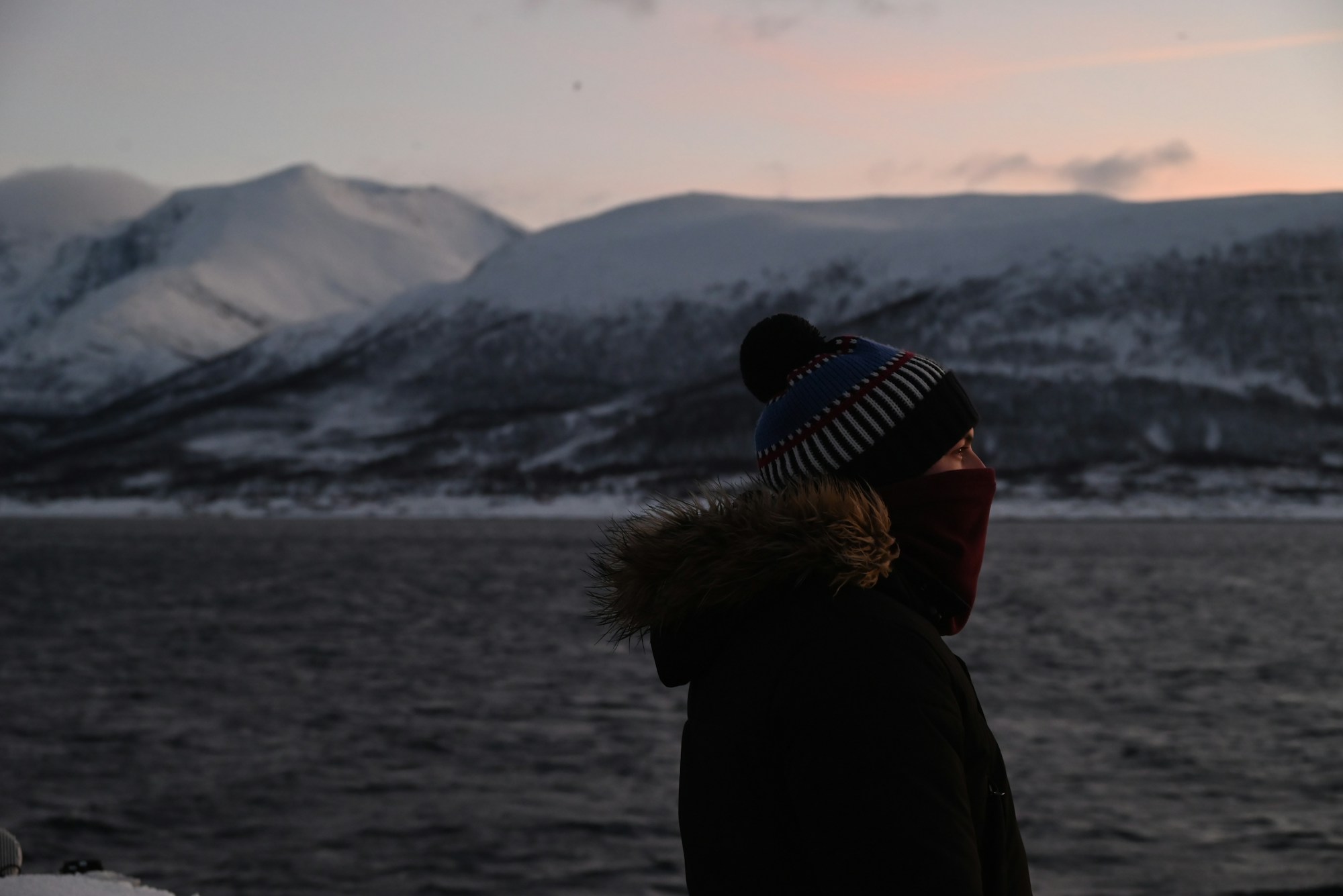 Reisender im Fellmantel mit Mütze vor einem verschneiten Fjord bei Tromsø, Norwegen