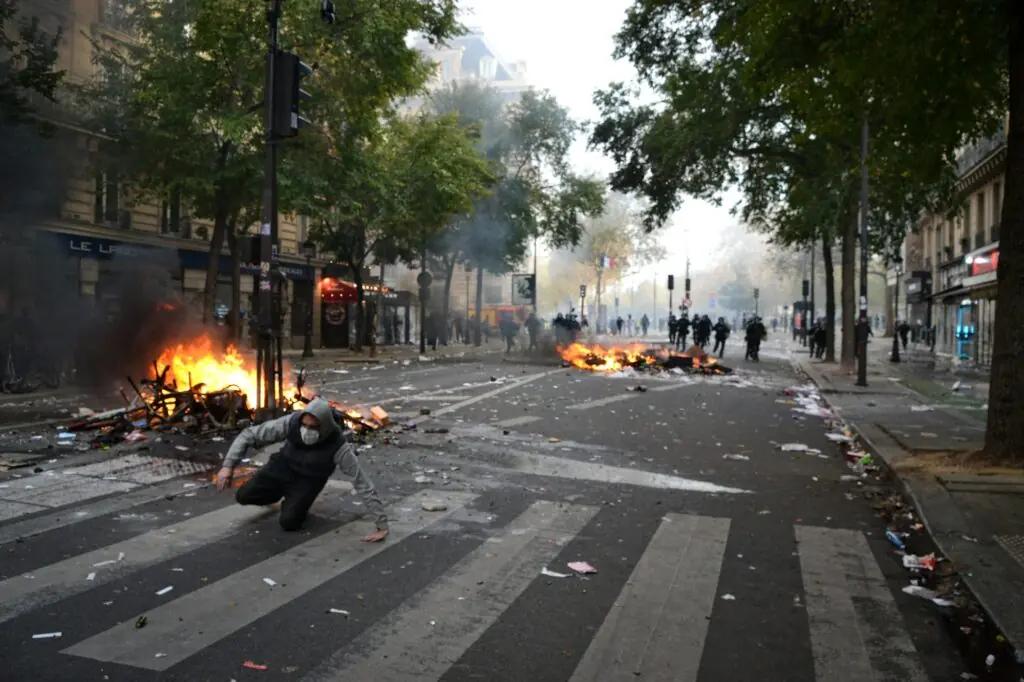 Personne encapuchonnée accroupie dans une rue de Paris, avec des débris en feu